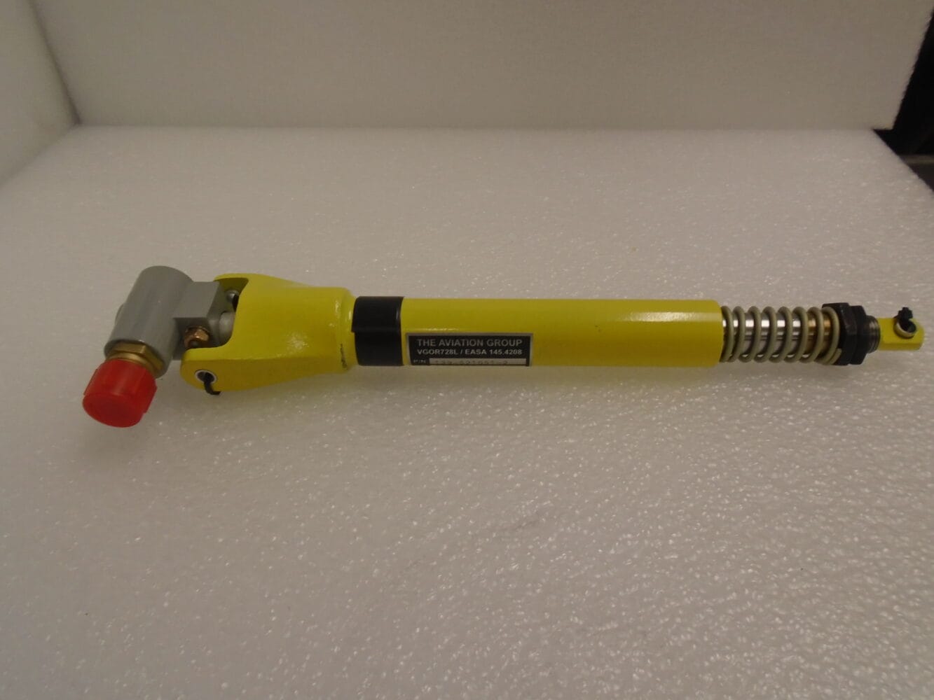 Yellow aircraft flap actuator with spring assembly and red protective cap, displayed on a white foam surface.